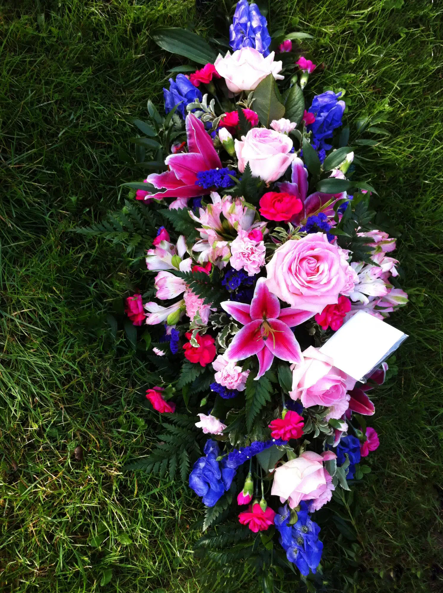 Colourful funeral flower arrangement with pink roses, lilies and blue flowers laid on grass